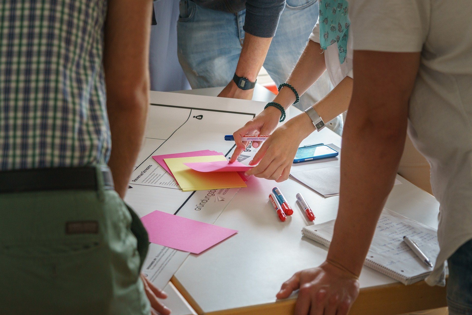 Eine Gruppe von Menschen steht um einen Tisch herum, der mit bunten Haftnotizen, Papieren, Stiften, einem Notizbuch und einem Smartphone bedeckt ist. Sie scheinen an einem Projekt zusammenzuarbeiten oder Ideen zu sammeln. Nur ihre Oberkörper und Hände sind sichtbar.