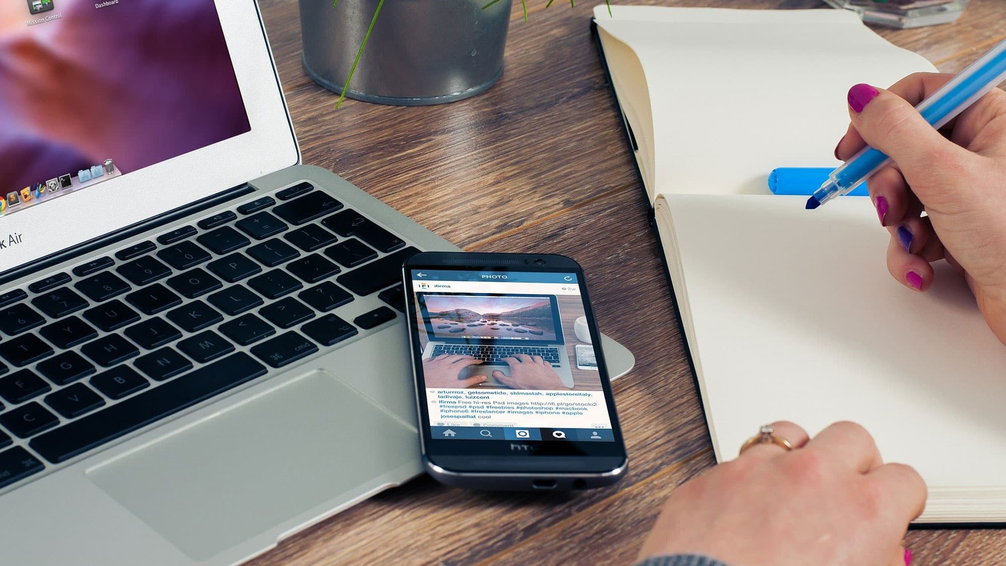 Eine Person schreibt mit einem blauen Stift in ein Notizbuch neben einem offenen Laptop und einem Smartphone, auf dem ein Instagram-Beitrag angezeigt wird. Auf dem Schreibtisch stehen außerdem eine Pflanze und ein kleiner Behälter mit Stiften. Der Arbeitsplatz wirkt organisiert und modern.