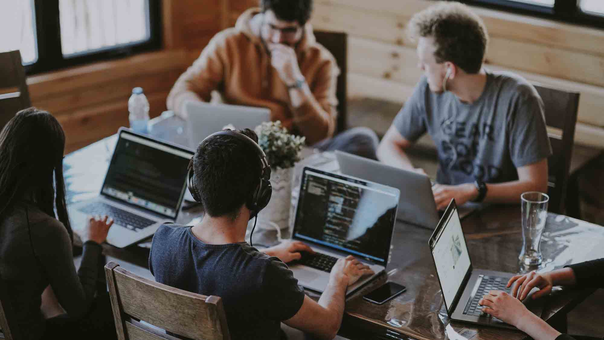 Eine Gruppe von fünf Personen sitzt um einen Tisch herum und arbeitet an Laptops. Auf dem Tisch steht eine Vase mit Blumen und anderen Gegenständen. Eine Person trägt Kopfhörer, während die anderen mit anderen Aktivitäten beschäftigt sind. Der Raum hat Holzwände und große Fenster.