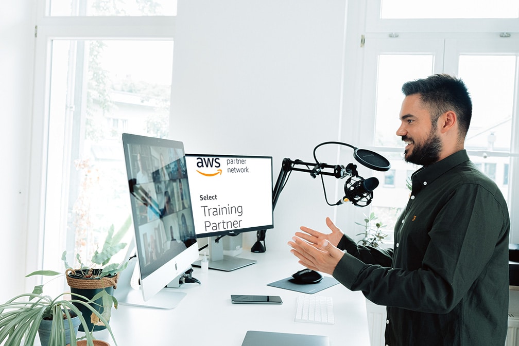 Ein Mann steht an einem Schreibtisch und spricht in ein Mikrofon mit Pop-Filter. Vor ihm steht ein Computerbildschirm, auf dem ein virtuelles Meeting und das AWS-Logo angezeigt werden. Es scheint, als würde er eine Präsentation halten oder eine Online-Schulung durchführen. Auf dem Schreibtisch stehen Pflanzen und eine Tastatur.
