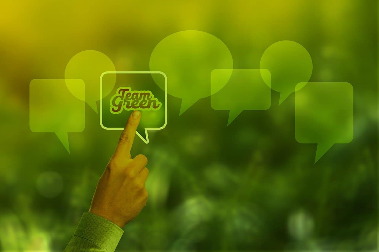 Eine Hand zeigt auf eine grüne Sprechblase mit dem Text „Team Green“ auf einem Hintergrund aus durchscheinenden grünen Sprechblasen und einem unscharfen natürlichen grünen Hintergrund. Das allgemeine Farbthema sind verschiedene Grüntöne.