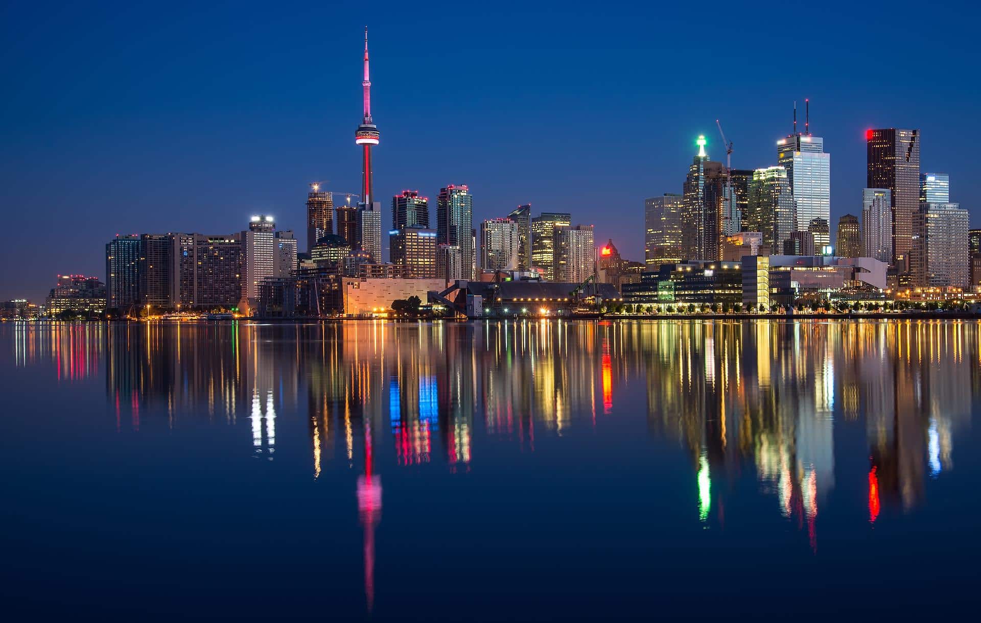 Eine nächtliche Stadtansicht von Toronto zeigt den beleuchteten CN Tower und verschiedene Hochhäuser. Die Lichter der Stadt erzeugen farbenfrohe Spiegelungen auf der ruhigen Uferpromenade und betonen die heitere Atmosphäre der Szene.