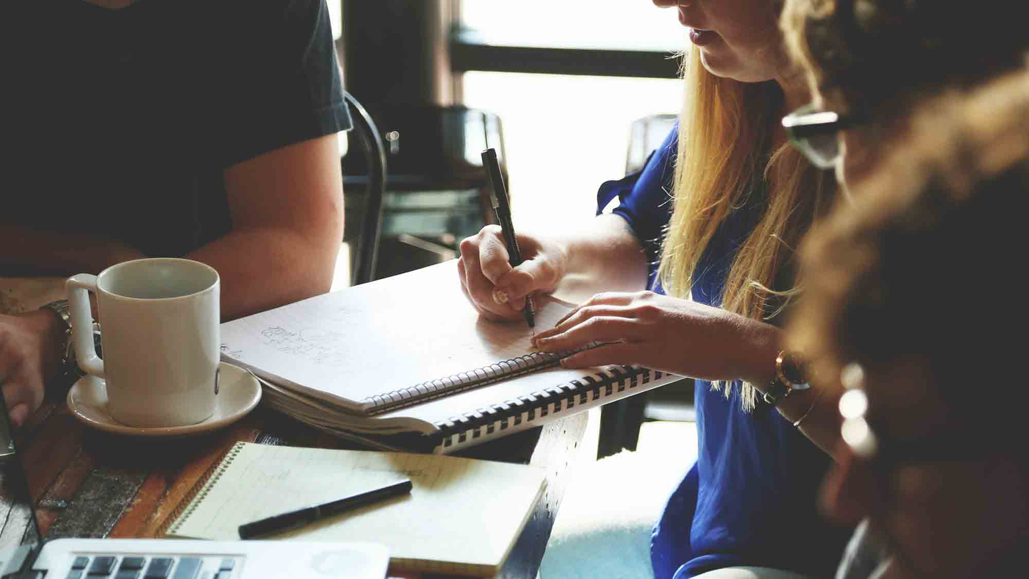 Eine Gruppe von Menschen sitzt um einen Holztisch, diskutiert und macht sich Notizen. Eine Person schreibt mit einem Stift in ein Notizbuch. Auf dem Tisch stehen Kaffeetassen, Notizbücher und ein Laptop, was auf ein gemeinsames Meeting oder eine Brainstorming-Sitzung schließen lässt.