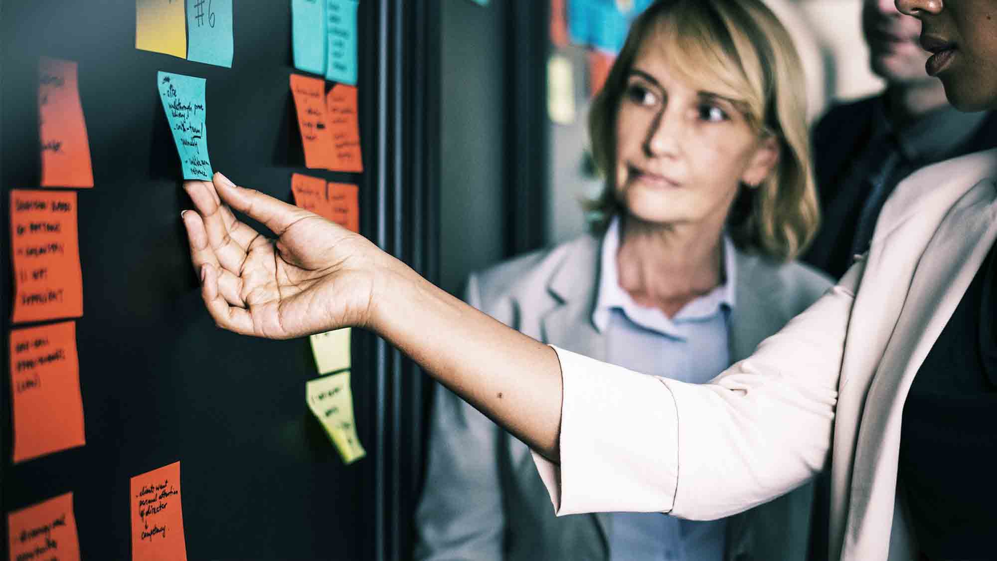 Eine Gruppe von Menschen steht vor einer Wand, die mit bunten Haftnotizen bedeckt ist. Eine Person streckt ihre Hand aus, um eine blaue Haftnotiz an die Wand zu kleben. Sie scheinen an einem Brainstorming oder einer Planungssitzung teilzunehmen.