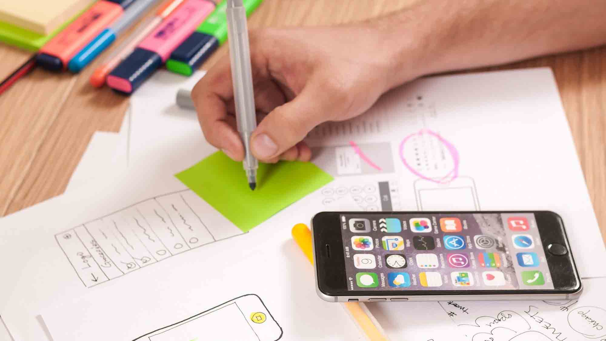 Man sieht die Hand einer Person, die einen Stift hält und auf einen grünen Haftnotizzettel schreibt, der auf einem Holzschreibtisch verstreut ist. Die Papiere scheinen Skizzen und Notizen zu enthalten. Neben der Hand liegt ein iPhone mit verschiedenen App-Symbolen. Im Hintergrund sind Textmarker und Marker zu sehen.