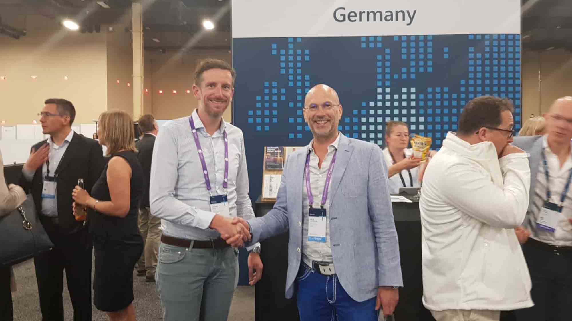 Zwei Männer schütteln sich vor einem Stand mit der Aufschrift „Deutschland“ bei einer Wirtschaftsveranstaltung die Hände. Beide lächeln und tragen Konferenzausweise. Im Hintergrund sind weitere Personen zu sehen, die sich unterhalten. Einer trägt einen hellen Blazer, der andere ein weißes Hemd und graue Hosen.