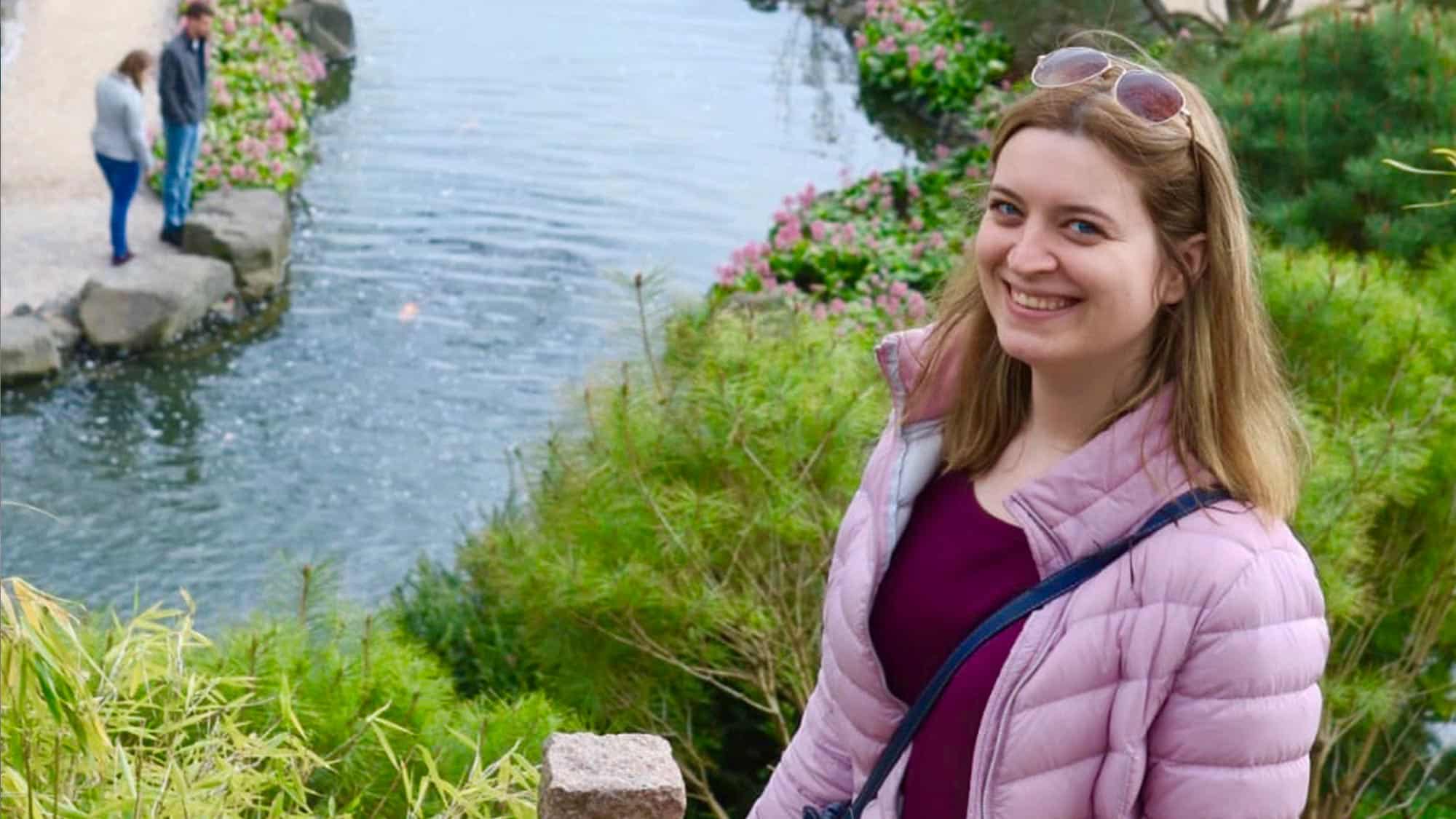 Eine Frau mit langen Haaren, einer hellvioletten Pufferjacke und einer Sonnenbrille auf dem Kopf lächelt in die Kamera. Sie steht neben einem lebhaften Garten mit einem Teich. Im Hintergrund bewundert ein Paar das Wasser und die umliegenden Blumen.
