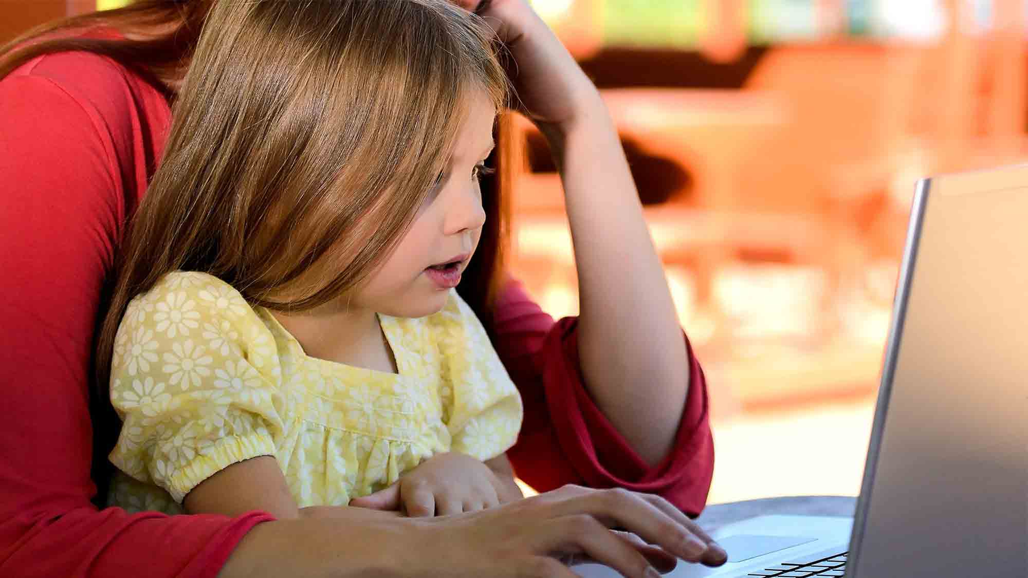Ein kleines Kind mit langen Haaren und einem gelben Kleid schaut fasziniert auf den Bildschirm eines Laptops, während ein Erwachsener in einem roten Hemd dicht hinter ihr sitzt und ihre Hände über die Tastatur führt. Der Hintergrund ist leicht verschwommen.