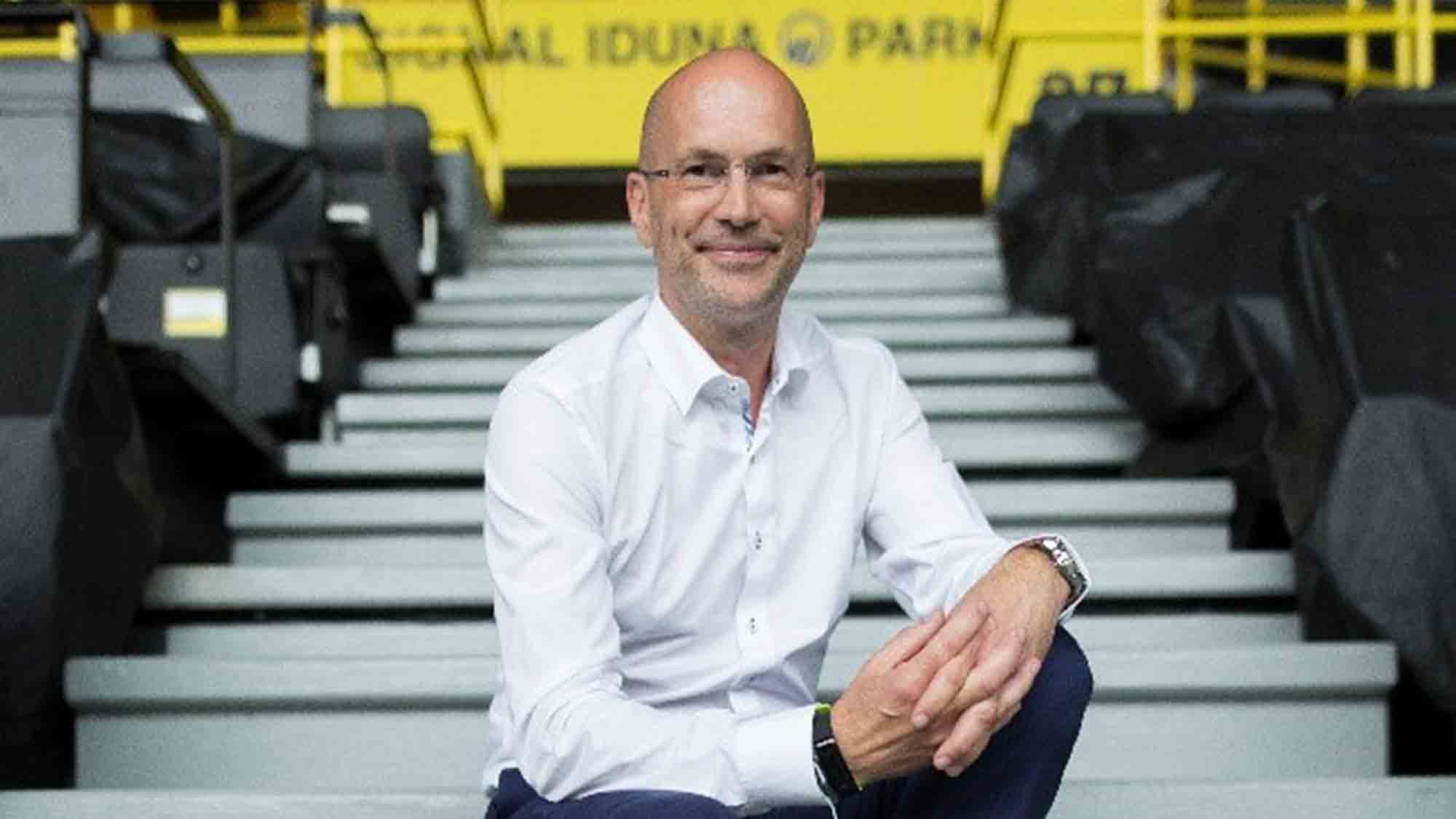 Ein Mann mit Brille und weißem Hemd sitzt auf Betonstufen in einem Stadion und lächelt in die Kamera. Im Hintergrund sind leuchtend gelbe Geländer und teilweise sichtbare schwarze Sitze zu sehen. Oben ist teilweise der Schriftzug „SIGNAL IDUNA PARK“ zu sehen.