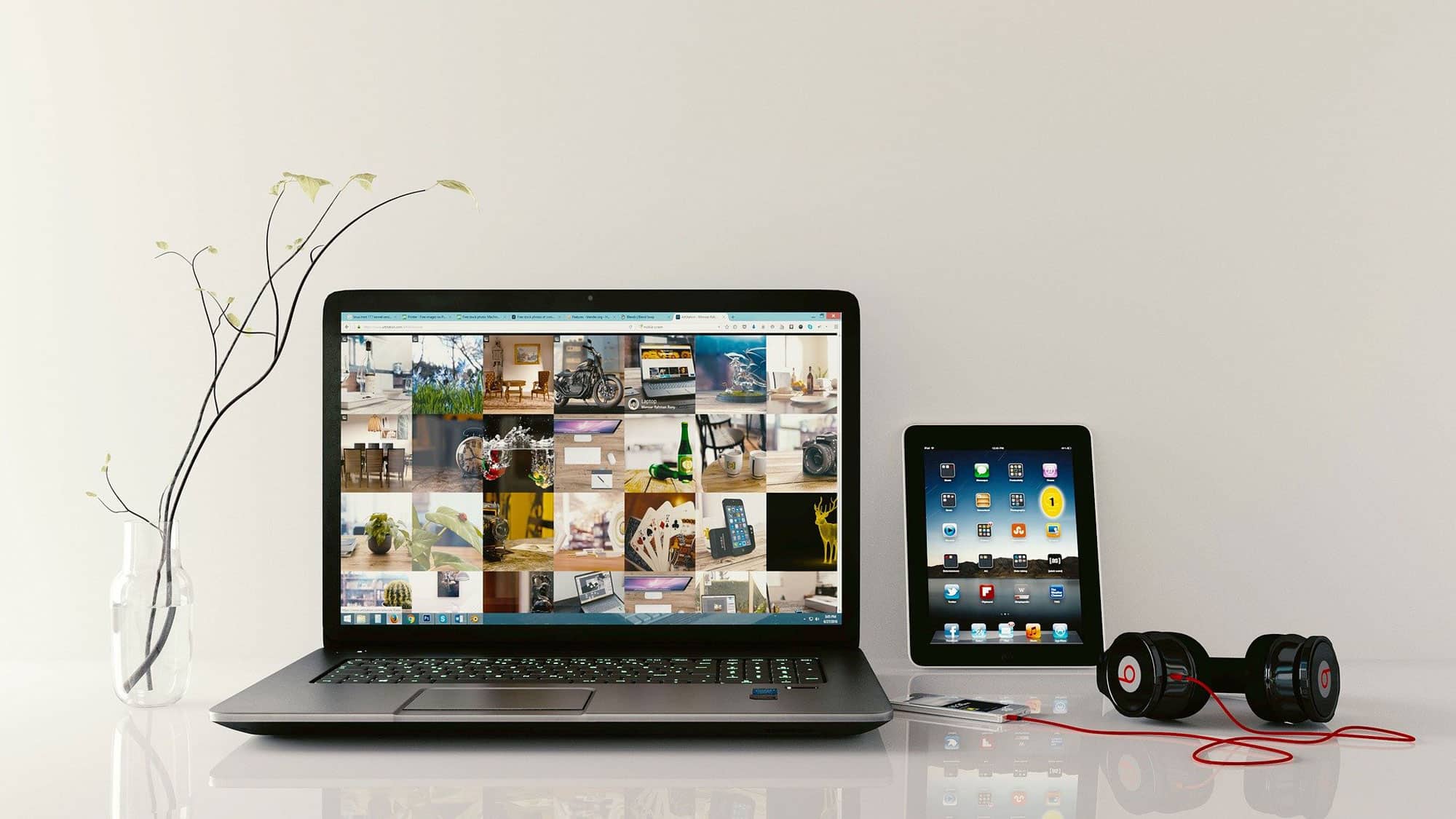 Ein sauberer Arbeitsplatz mit einem Laptop, auf dem verschiedene Bilder angezeigt werden, einem Tablet mit App-Symbolen, roten und schwarzen Kopfhörern, einem MP3-Player und einer minimalistischen Glasvase mit einem Zweig, der kleine weiße Blumen hält. Der Hintergrund ist eine schlichte, helle Wand.