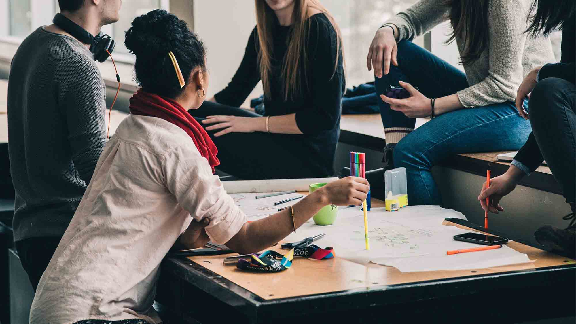 Eine Gruppe von fünf Personen versammelt sich um einen Tisch, der mit Papieren, Stiften und einer Tasse bedeckt ist. Eine Person mit einem Bleistift im Haar zeigt auf das Papier. Die anderen sitzen entspannt da und unterhalten sich. Sonnenlicht fällt durch die nahen Fenster und schafft eine helle Atmosphäre.