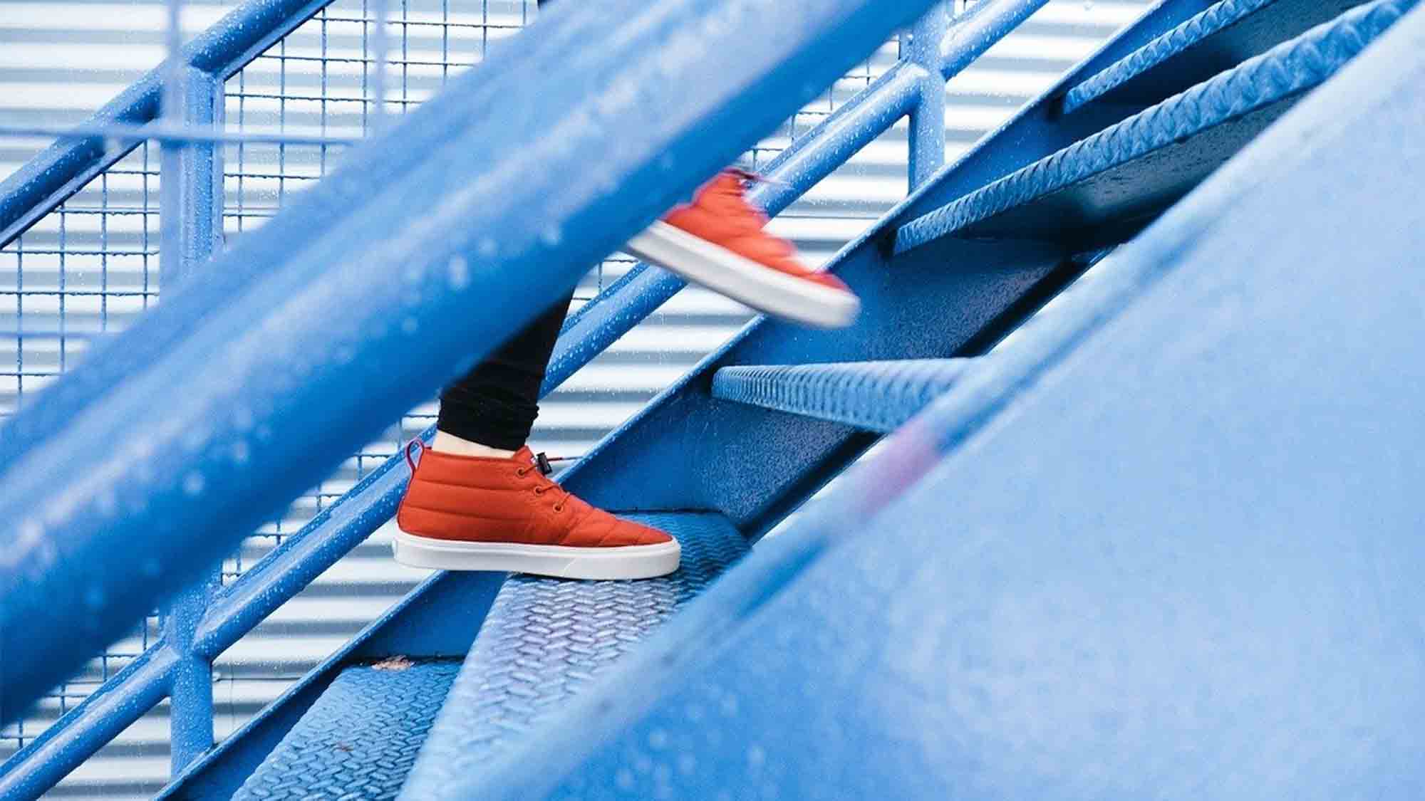 Eine Person in roten Turnschuhen geht eine hellblaue Metalltreppe hinauf. Das Foto ist schräg aufgenommen und fokussiert auf die Beine und Füße der Person. Im Hintergrund ist eine verschwommene, eingezäunte Struktur zu sehen.