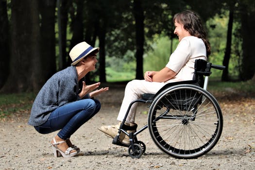 Zwei Frauen im Freien, eine steht, trägt einen Hut und hockt, während sie lächelt und mit der anderen Frau spricht, die im Rollstuhl sitzt. Sie stehen auf einem von Bäumen umgebenen Kiesweg und unterhalten sich fröhlich.