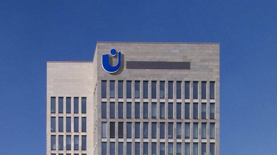 Ein modernes Hochhaus mit einem Raster aus gleichförmigen Fenstern vor einem klaren blauen Himmel. Das Gebäude verfügt oben über ein blau-weißes Logo, das aus einer stilisierten U-Form mit einem Punkt darin besteht.